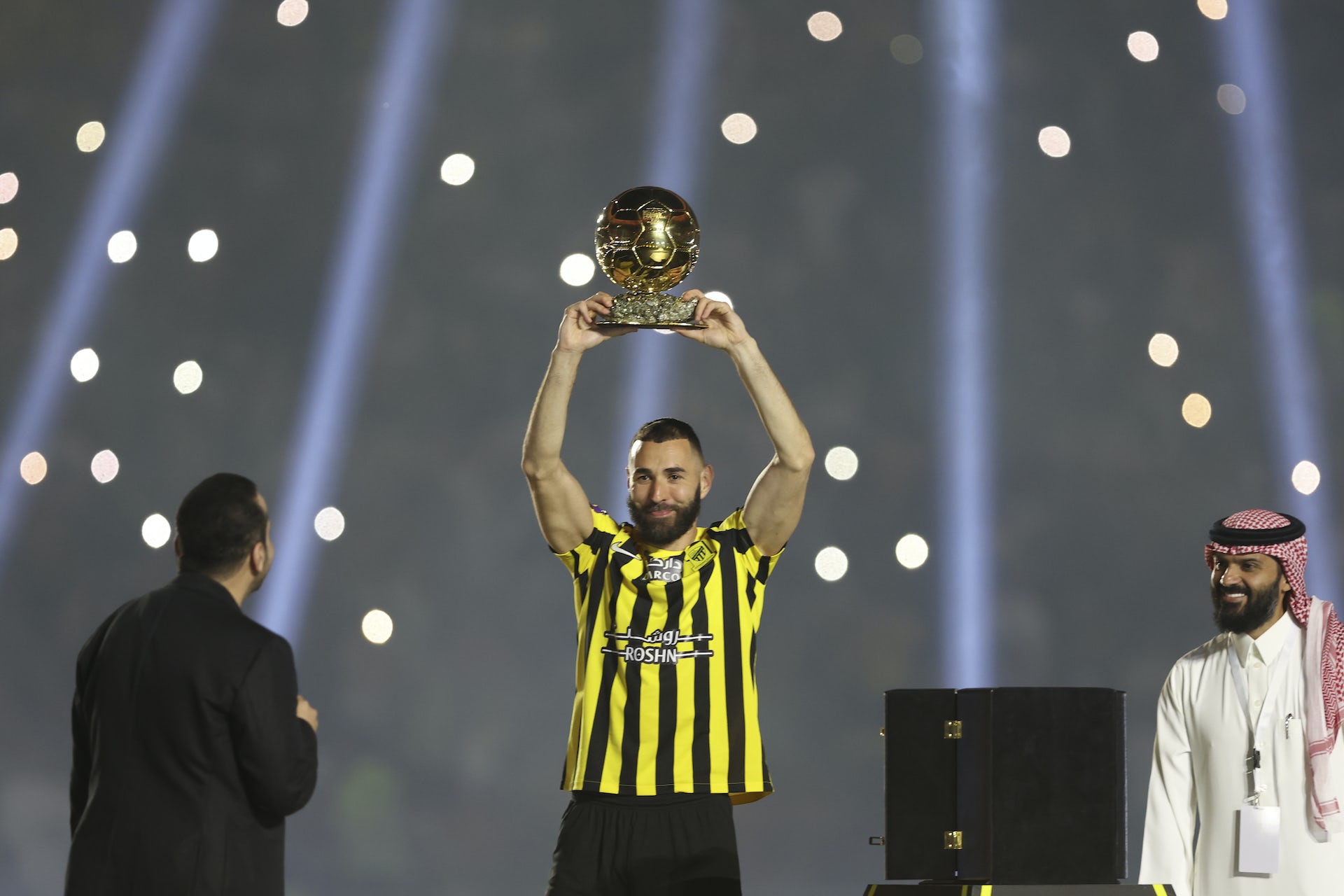 Un barbat intr-un tricou de fotbal negru si galben isi ridica un trofeu de aur deasupra capului.